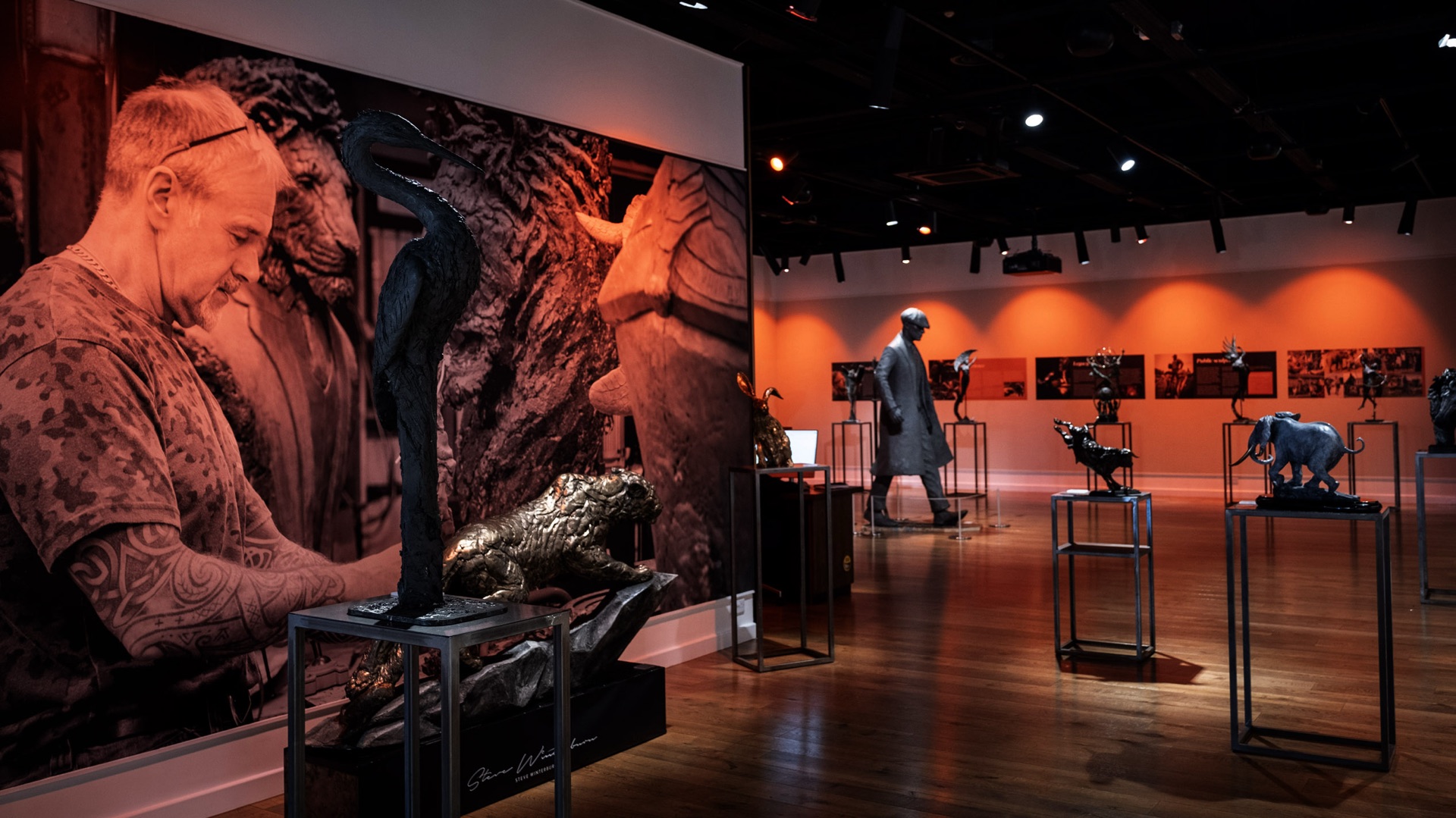 Bronze animal sculptures on plinths in a gallery lit in orange, beside a large print of the sculptor at work