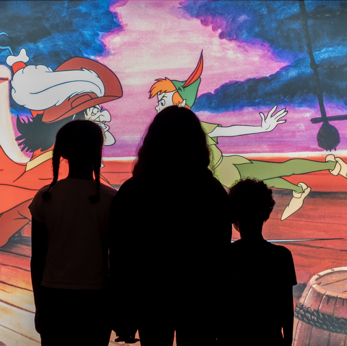 Three children in silhouette watching a projected Peter Pan and Captain Hook scene