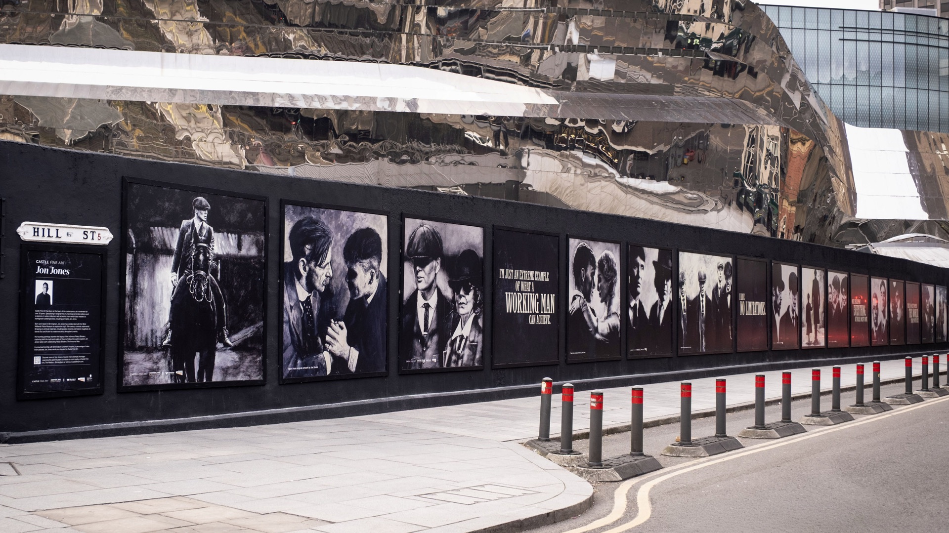 Street hoarding lined with black and white Peaky Blinders-style artwork prints beneath a mirrored building