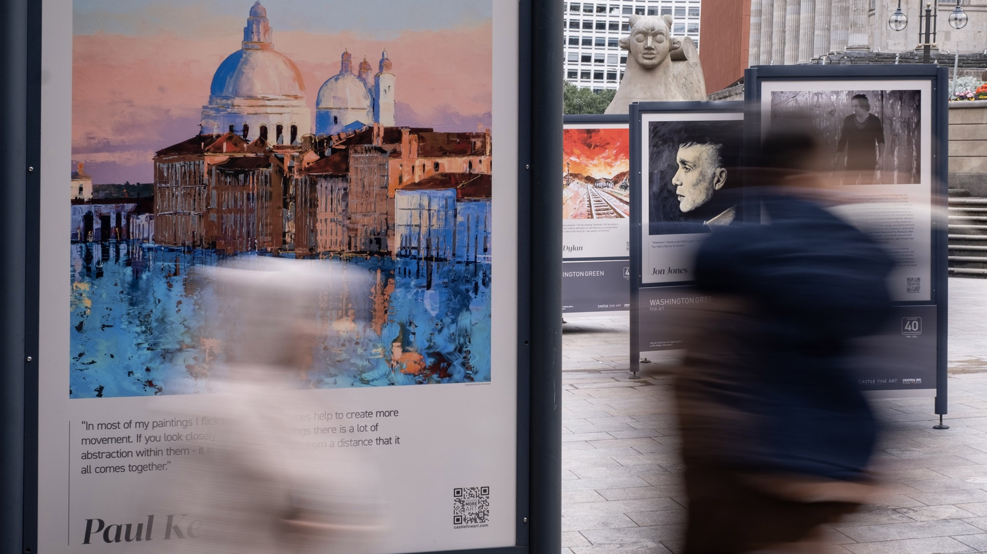 Outdoor art exhibition boards in a city square, with passers-by blurred in motion in front of a Venice painting