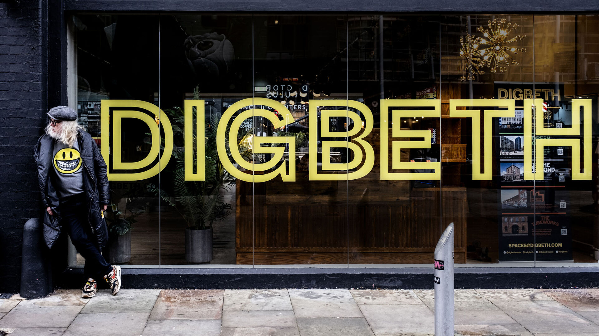 A figure in a smiley-face jacket outside a Digbeth shopfront lit by a yellow neon sign