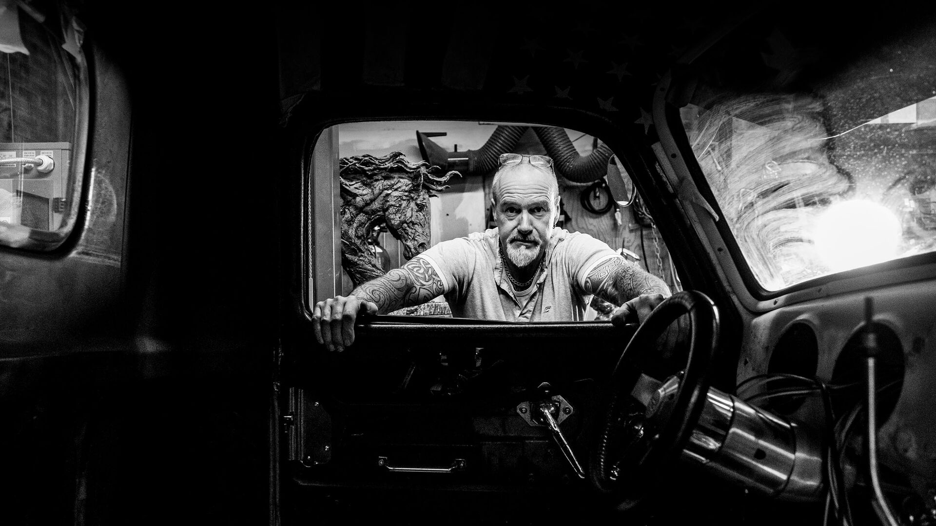 A black-and-white portrait of a tattooed man at the wheel of a vintage truck